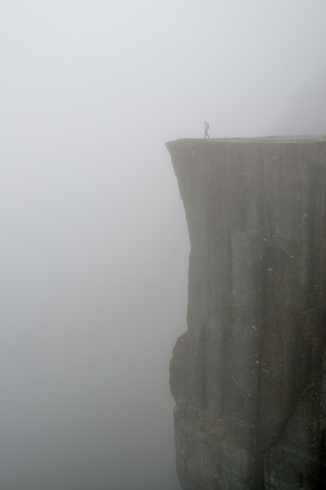 A figure wearing a backpack walks towards a vertical cliff edge in the fog. Photo by Anastasia Saldatava on Unsplash