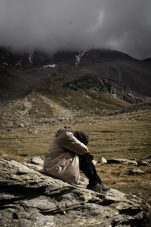 Exhausted and despondent looking person sat on a rock in a stark mountainous landscape with ominous looking weather closing in. Photo by Germán Rodríguez on Unsplash