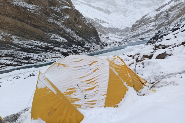 Yellow tent partially covered in snow on a snowy mountainside, beside a frozen river. Rugged cliffs in the background convey a remote, cold atmosphere. Photo by Joshi Milestoner on Unsplash