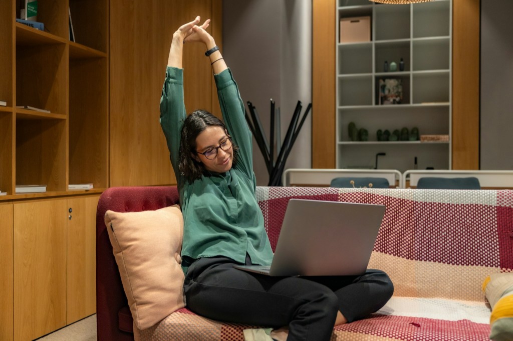 Woman stretches with arms overhead while seated comfortably on a patterned sofa, laptop on her lap, creating a cozy, relaxed scene. Photo by Resume Genius on Unsplash.